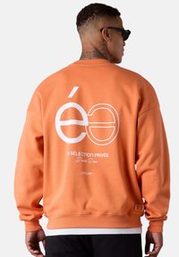 Sweatshirt orange oversize avec un logo blanc au dos. Doté de poignets et d'ourlet côtelés, fabriqué en tissu doux avec une texture lisse.