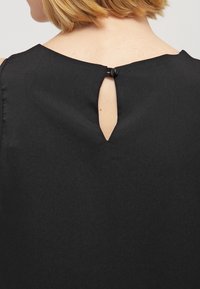 Top negro sin mangas que presenta un pequeño corte en forma de llave en la parte posterior, asegurado con un botón negro, textura de tela suave.