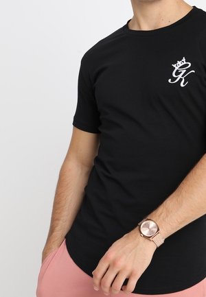 Mies, jolla on musta t-paita, jossa valkoinen "GK"-logo, ja ruusukultainen rannekello, käsi vaaleanpunaisen housun taskussa, yksivärinen vaalea tausta.