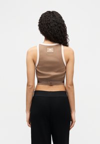 Γυναίκα με σγουρά μαλλιά, που φοράει μπεζ κοντό τοπ με λογότυπο στην πλάτη και μαύρο ψηλόμεσο παντελόνι, στέκεται μπροστά σε λευκό φόντο.