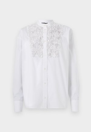 Chemise blanche à boutons avec un détail floral texturé sur la poitrine, des manches longues et un col Mao. Tissu en coton lisse.