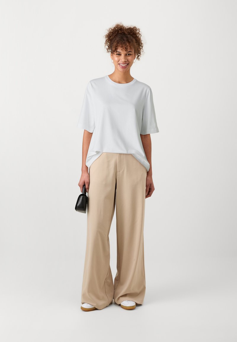Vit t-shirt med kort ärm och avslappnad passform, kombinerad med beige vida byxor. Modellen håller en liten svart handväska och har på sig vita sneakers.