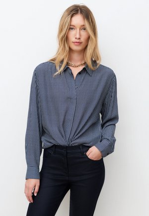 Vaaleatukkainen nainen, joka pukeutunut kierrekuidulla kuvioituun laivastonsiniseen ja valkoiseen ruutupaitaan, mustiin housuihin ja kultaketjuun, seisomassa valkoista taustaa vasten.