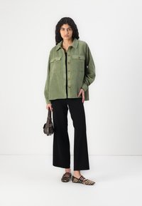 Green corduroy shirt jacket with two front pockets, black top underneath, wide-leg black pants, and leopard print flats. Carrying a small handbag.