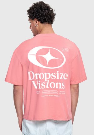 Man with curly black hair and earrings wearing a loose pink t-shirt with white graphic text and symbols on the back.