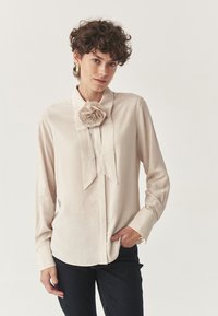 Blusa beige chiaro con maniche lunghe, colletto e un grande dettaglio floreale sul colletto. Tessuto morbido e vestibilità sagomata.