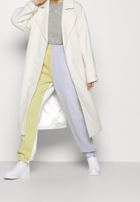 Vit lång kappa, grå t-shirt och pastellfärgade sweatpants (gul och ljuslila) med vita sneakers, som visar en avslappnad passform och lagerdesign.