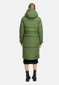 Kvinna som står med ryggen vänd, iklädd en lång olivgrön vadderad kappa med huva, svart kjol och svarta ankelstövlar.