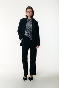 Žena stojí oblečená v čiernom saku, čiernych nohaviciach, trblietavom striebornom tope a čiernych lodičkách s veľkými mašľami, na pozadí bielej steny.