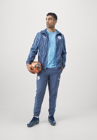 Tuta atletica blu e navy con giacca a fantasia, maglietta a maniche corte e pantaloni. Tiene in mano un pallone da calcio arancione e nero.