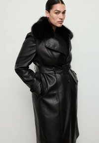 Cappotto in pelle nera con collo in pelliccia sintetica, vita con cintura, ampie rever e tasche laterali. Texture liscia con una vestibilità su misura.