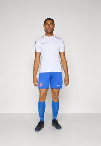 Hvid kortærmet atletisk skjorte med sorte detaljer, parret med blå fodboldshorts med et logo, blå knæstrømper og sorte fodboldstøvler.