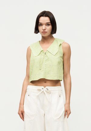 Jonge vrouw met kort donker haar, die een mouwloos lichtgroen geruit cropped top en witte broek met trekkoord draagt, tegen een effen achtergrond.