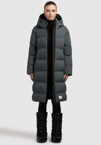 Lang donsjack in donkergrijs, met capuchon, rits aan de voorkant en gewatteerd ontwerp. Vergezeld van zwarte, zware winterlaarzen.
