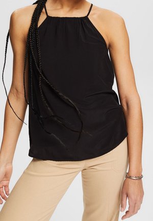 Frau trägt ein ärmelloses schwarzes Neckholder-Top und beige Hosen, mit lang geflochtenem Haar und einem silbernen Armband am rechten Handgelenk.