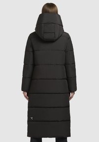 Moteris atrodo nuo nugaros, vilkinti ilgą juodą siūlometį pūkinį palto su dideliu kapišonu, priešais vientisą pilką foną.