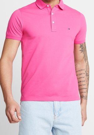 Mann trägt ein hellpinkes kurzärmeliges Polohemd und hellblaue Jeans, mit einem Schriftzug-Tattoo am rechten Unterarm und einem Teil des linken Arms sichtbar.