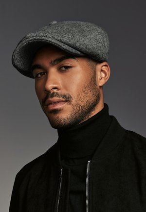 Gray wool flat cap with a rounded top and a small button accent, worn over a black turtleneck and jacket, set against a neutral background.