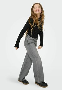 Juoda siaurinta ilgomis rankovėmis viršūnė derinama su laisvomis pilkomis plataus silueto džinsinėmis kelnėmis. Juodi sportbačiai su ruda pado dalimi. Žaisminga poza prieš šviesią foną.