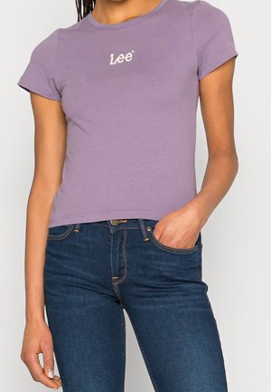 Mladá osoba nosí přiléhavé levandulově fialové tričko Lee a tmavě modré džíny s jednou rukou v kapse na jednobarevném pozadí.