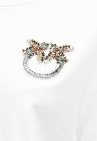 Διακοσμητικό καρφίτσα σε σχήμα φιόγκου με πολύχρωμους λίθους σε διαφανή, κεχριμπαρένια, τυρκουάζ και ελιά απόχρωση σε λευκό υφασμάτινο φόντο.