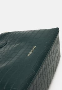 Zelená kabelka z umělé krokodýlí kůže s obdélníkovým tvarem, hladkou strukturou a zlatým logem "Valentino" na přední straně.