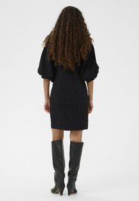 Vestido negro texturizado con mangas cortas abullonadas, dobladillo a la altura de la rodilla, combinado con botas de cuero negro ajustadas, hasta la rodilla, y tacones afilados.
