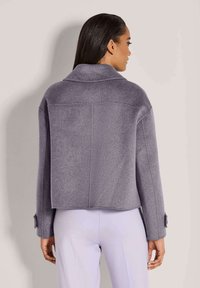 Frau mit langen dunklen Haaren, die eine graue, langärmlige Cropped-Jacke und helllavendelfarbene Hosen trägt, vor einem schlichten Hintergrund stehend.