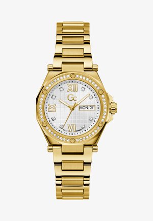 Kuldtooni kellok, millel on oktaadiline korpus, valge tekstureeritud kellanägu, rooma numbrid, kuupäeva näidik ning kristallaktsentidega bezel ja kellanägu. Metallist ketiga rihm.