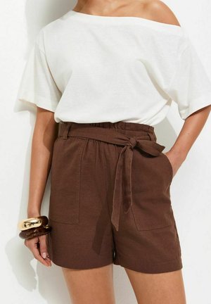 Kobieta nosząca biały top z odkrytymi ramionami i wysokim stanem brązowe szorty z paskiem zawiązanym w kokardę, uzupełnione masywnymi bransoletkami na lewym nadgarstku.