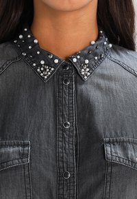 Grey denim shirt featuring a studded collar with pearls and metallic accents, front button closure, and a single chest pocket.
