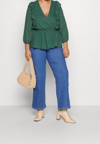 Femme portant un chemisier vert à volants, un jean bleu, des bottines beiges à lacets, tenant un sac à main beige texturé sur un fond uni.