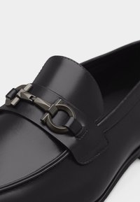 Mocassin en cuir noir à finition polie, avec un détail boucle mors métallique sur le dessus.