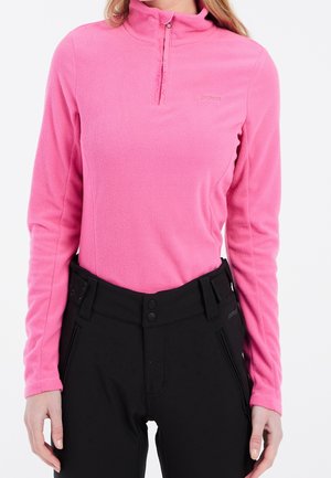 Frau trägt ein hellpinkes Fleecepullover mit langen Ärmeln, Viertel-Reißverschluss und schwarze hochtaillierte Hose mit Knopfverschluss.