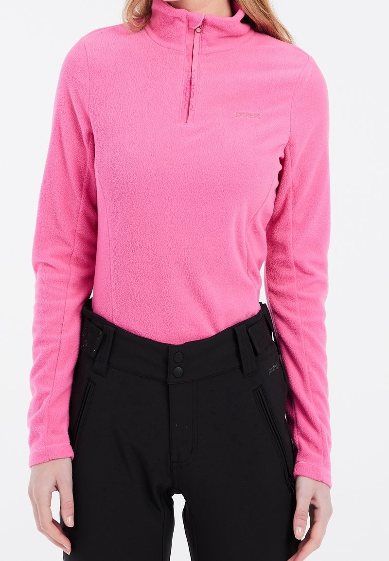 Femme portant un haut polaire à manches longues rose vif avec une fermeture éclair quart et un pantalon noir taille haute avec fermeture à boutons.