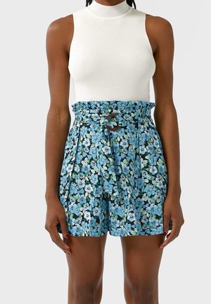 Haut blanc sans manches en côte, associé à un short bleu fleuri taille haute avec une taille paperbag et une ceinture nouée avec une boucle circulaire.