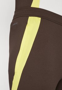Leggings de sport marron avec un panneau latéral en maille jaune, texture de tissu lisse et un petit logo "AEROREADY" sur la taille.