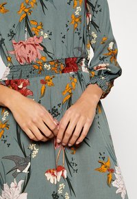 Abito floreale in teal smorzato, con fiori rossi, rosa e arancioni. Vita e polsini arricciati, tessuto strutturato e sottili accenti di uccelli.