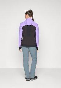 Giacca nera e viola con cappuccio, caratterizzata da un pannello posteriore texturizzato. Abbinata a pantaloni blu chiaro e scarpe da escursionismo nere.