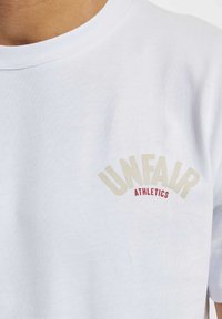 Hvit bomull t-skjorte med beige og rød "UNFAIR ATHLETICS" grafikk på brystet, med klassisk rund hals og korte ermer.