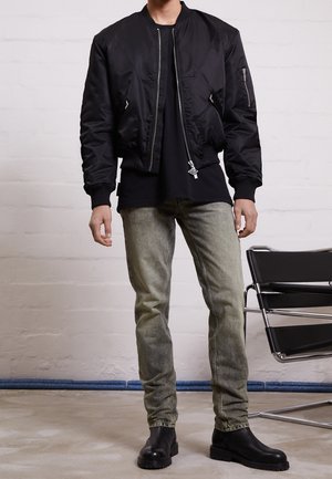 Personne portant une veste bomber noire, une chemise noire, un jean gris délavé et des bottes noires, debout à côté d'une chaise en métal noir et cuir.