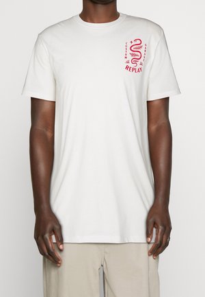 Homme portant un t-shirt blanc uni à manches courtes avec un graphisme de serpent rouge et le texte « Custom Garage Replay » sur la partie supérieure gauche de la poitrine.