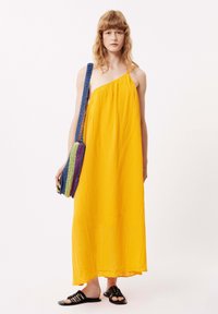 Gelbes Maxikleid mit einem strukturierten Muster, einem einseitigen Schulterträger, kombiniert mit einer bunten, gewebten Tasche und schwarzen, breiten Sandalen.