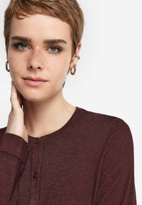 Burgundy stickad kofta med rund halsringning, med knäppning och en mjuk textur. Modellen framhäver plaggets passform.