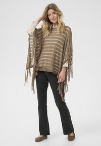 Poncho marron à franges avec un motif ajouré et des rayures horizontales, superposé sur un haut crème, associé à un jean noir évasé et des ballerines marron.