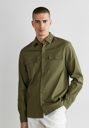 Mand iført en olivengrøn tekstureret skjorte med knapper og to brystlommer, stående mod en ensfarvet lys grå baggrund.