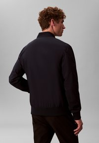 Veste bomber noire avec une texture lisse et mate, poignets et col côtelés. Design simple sans logos ni motifs visibles.