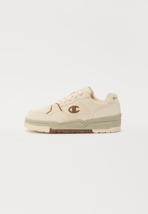 Beige sneaker met een gestructureerde canvas bovenkant, contrasterend bruin logo en stevige crèmekleurige en olijfgroene zool. Afgeronde neus en vetersluiting.