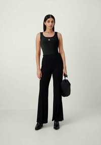 Black bodysuit with a square neckline and logo, paired with high-waisted, flared black trousers. Model holds a black, gathered handbag.