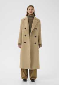 Cappotto beige doppiopetto con grandi bottoni neri, revers a farfallino e maniche lunghe, indossato sopra un maglione marrone a collo alto e pantaloni larghi.